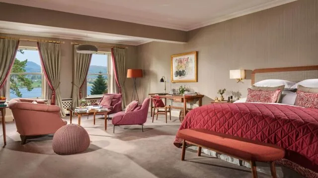 Spacious hotel room with a large bed, seating area, desk, and two windows overlooking trees and a lake; decor features pink and red tones.