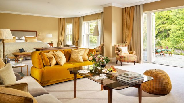 Spacious hotel room with a bed, yellow sofa, armchairs, glass coffee table with books and flowers, large windows, and open doors leading to a garden patio.