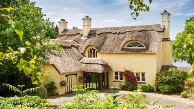 A yellow house with a thatched roof, chimney stacks, arched windows, and flowering plants in front, surrounded by greenery on a sunny day.