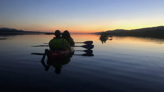 Serene sunset kayaking experience, guests paddle in calm waters with scenic mountain views, ideal for nature lovers.