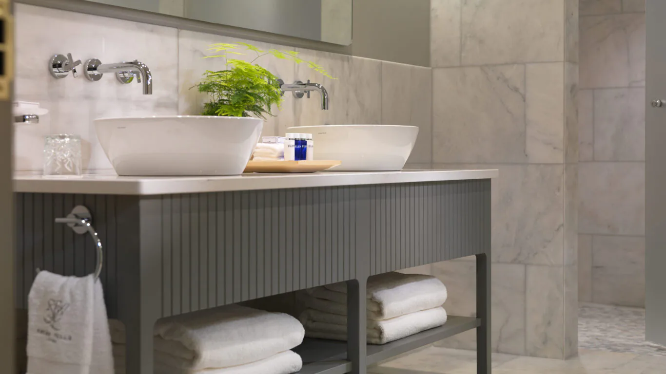 Elegant hotel bathroom with marble walls, twin basins, soft towels, a lush potted plant, and spa-like amenities.