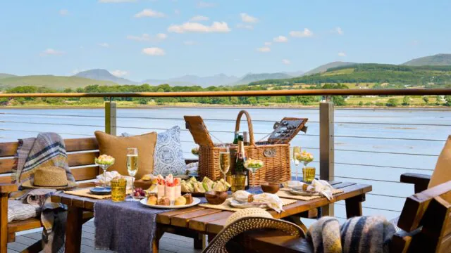 Scenic lake-view terrace with picnic, wine, and fresh snacks on a wooden table, perfect for relaxation and leisure.