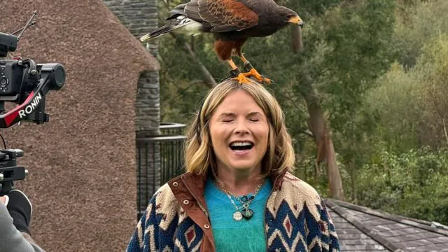 Guest laughs with a hawk perched on her head at a countryside hotel experience, surrounded by lush greenery, filmed for memories.