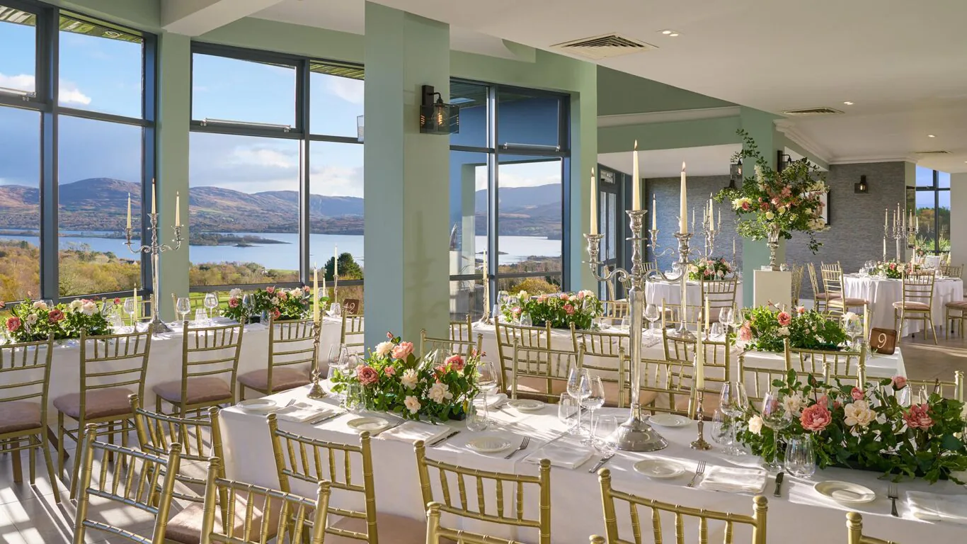 Elegant hotel dining room with panoramic lake views, adorned with floral arrangements and gold chairs, perfect for events.