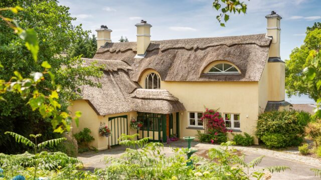 A yellow house with a thatched roof, arched windows, and a garden with flowers and greenery on a sunny day.