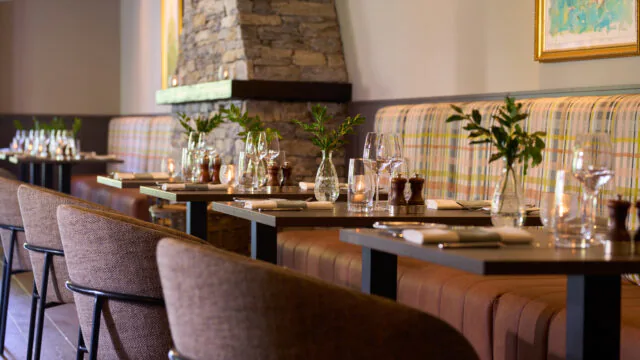 Cozy hotel dining area with elegant stone fireplace, plaid seating, set tables, and warm lighting, perfect for relaxing meals.