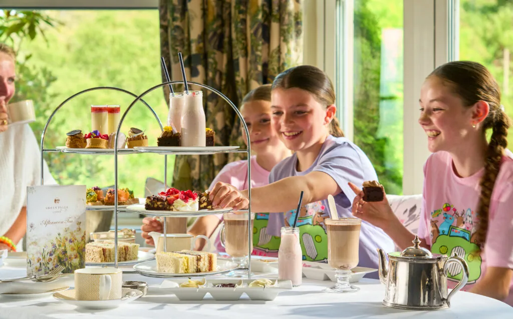 Guests enjoying elegant afternoon tea with pastries, sandwiches, and drinks, in a sunlit room with garden views.