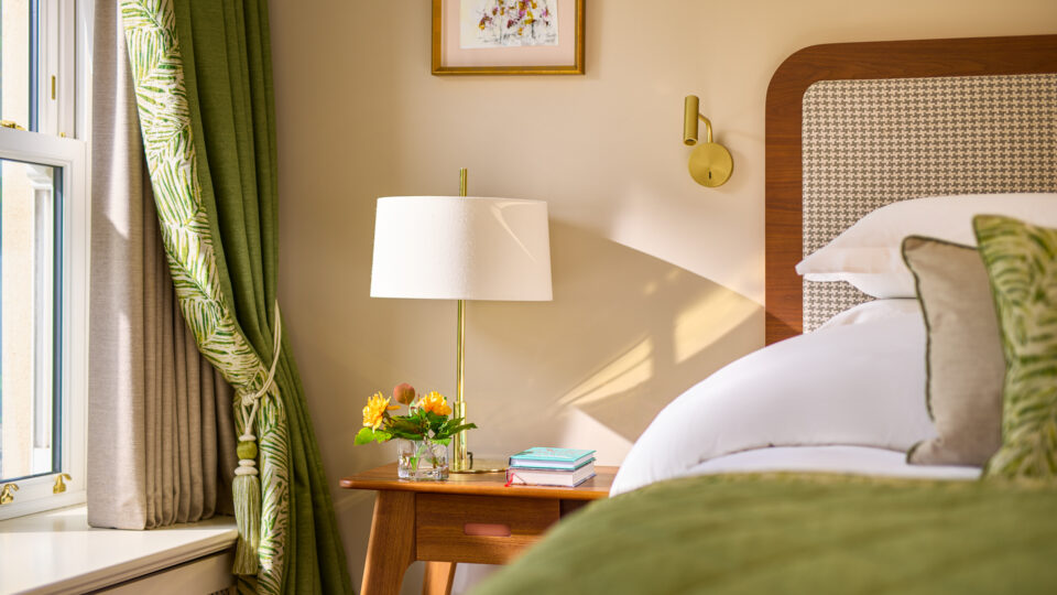 Elegant hotel room with plush bedding, green curtains, modern lamp, and fresh flowers offering a cosy, relaxing stay.