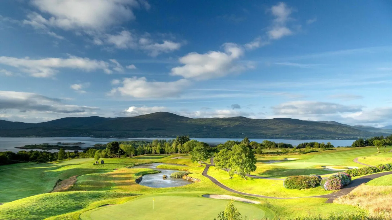Scenic view of golf course by a serene lake and rolling hills under a bright blue sky, perfect for a relaxing holiday.