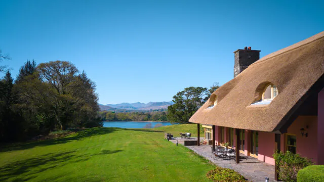 Charming hotel with thatched roof overlooking serene lake and mountains, lush green lawn, outdoor seating for relaxation.