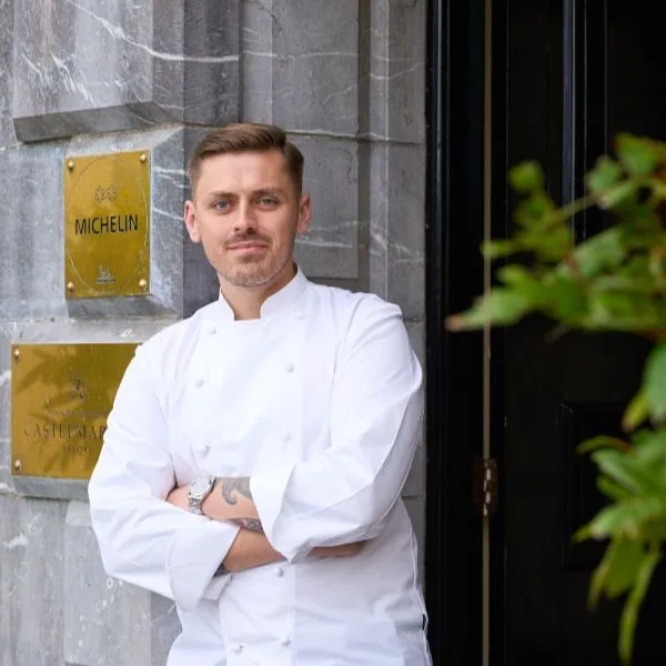 A chef in a white uniform stands with arms crossed in front of a building with a Michelin plaque on the wall.