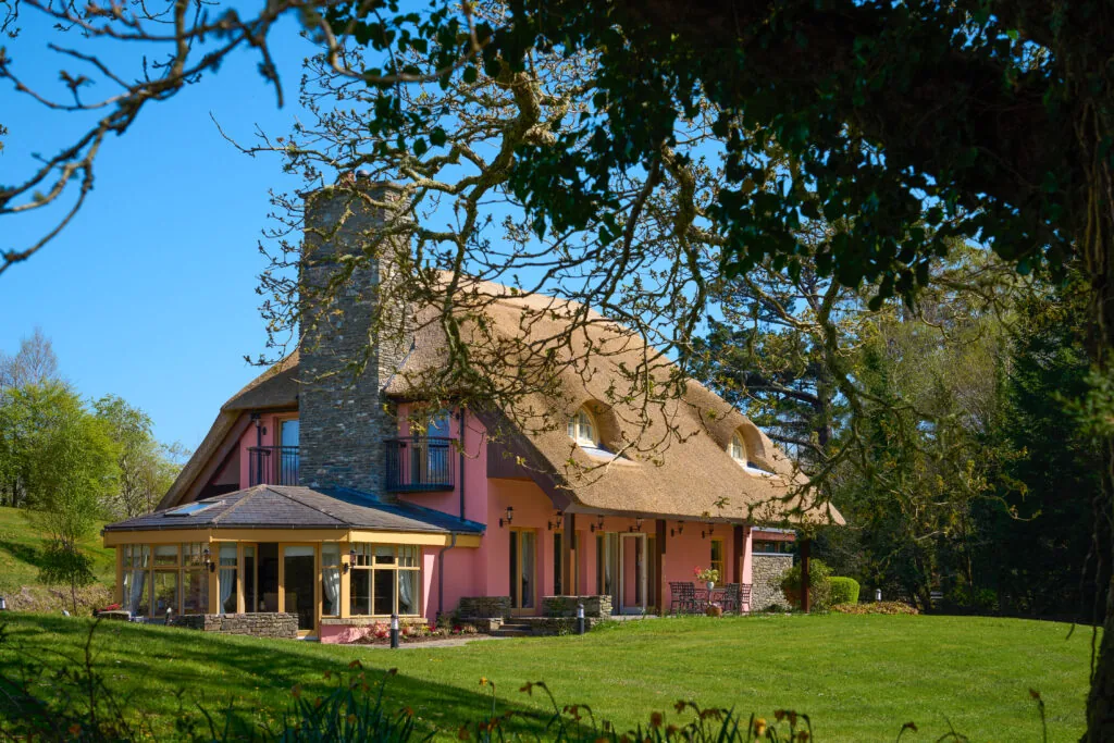 A pink two-story house with a thatched roof and large chimney sits on a grassy lawn, surrounded by trees and blue sky.