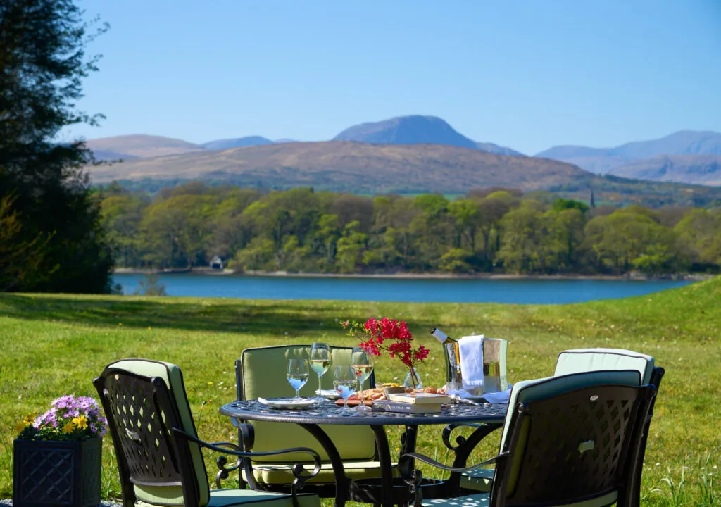 Scenic outdoor dining with mountain and lake views, featuring elegant table setting, wine, and lush greenery.