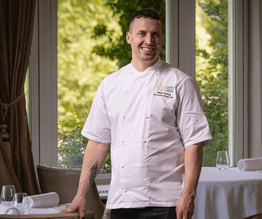 A chef in a white uniform stands in a restaurant dining area with set tables, smiling in front of large windows showing greenery outside.