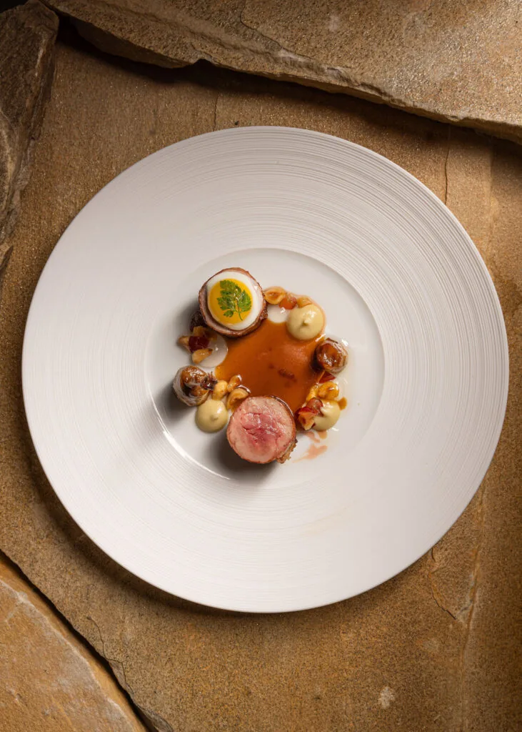 A white plate with two pieces of cooked meat, a halved stuffed egg, sauce, dollops of cream, and small garnishes, set on a stone surface.