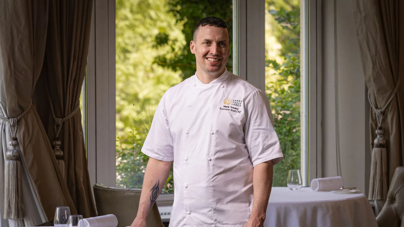 A chef in a white uniform stands and smiles in an elegant dining room with large windows and set tables.