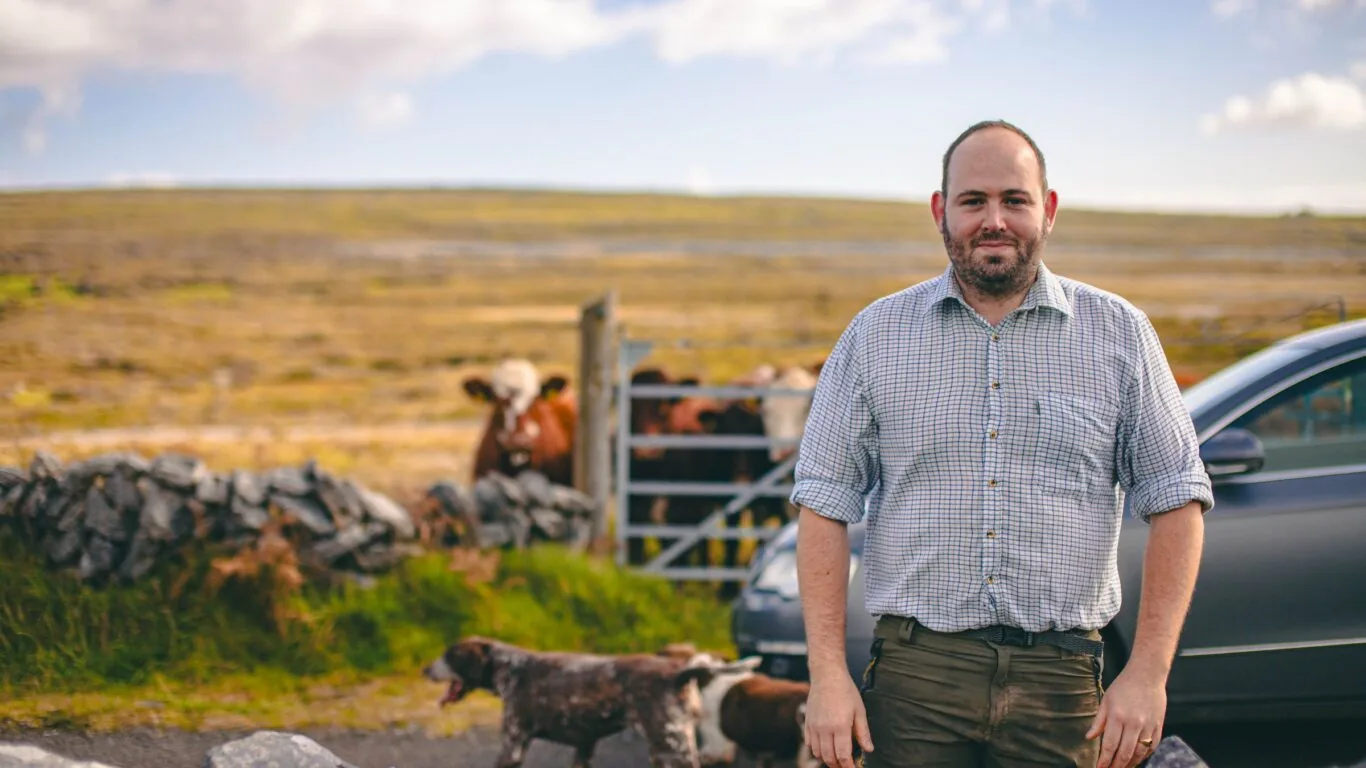 A man stands in front of a stone wall and parked car, with cows and a dog behind him in a rural landscape under a partly cloudy sky.