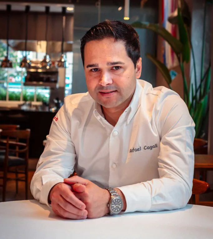 A man in a white chef&rsquo;s jacket sits at a table in a restaurant, facing the camera with hands clasped.