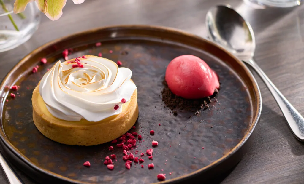 A dessert plate with a lemon meringue tart garnished with crushed raspberries and a scoop of bright pink sorbet on chocolate crumbs, alongside a spoon.