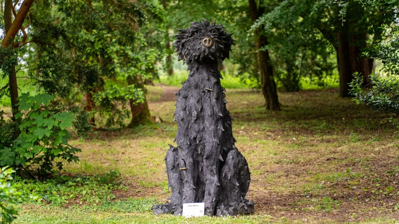 A large, textured sculpture of a seated dog with a shaggy coat stands on grass in a wooded park area. A small sign is placed in front of the statue.