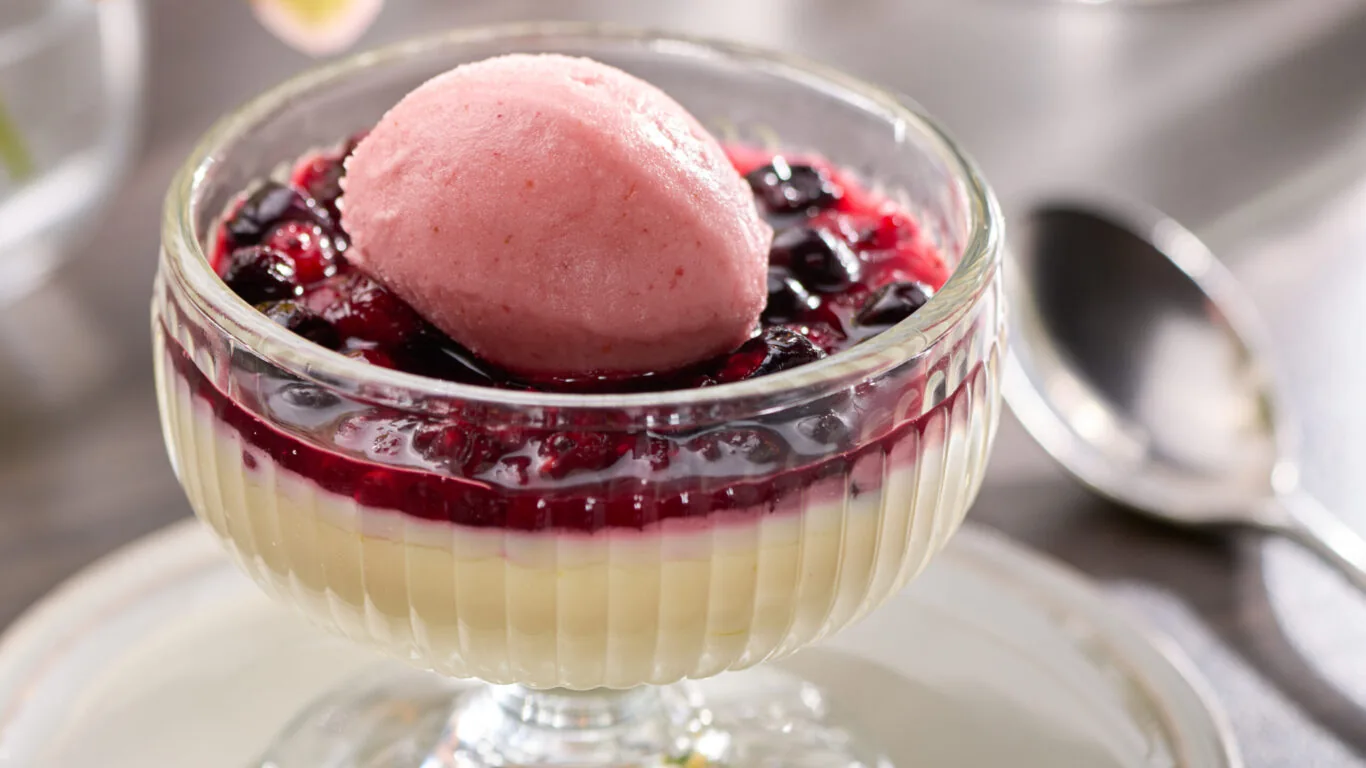 A glass dessert dish with creamy custard, topped with mixed berry sauce and a scoop of pink sorbet, sits on a saucer next to a spoon.
