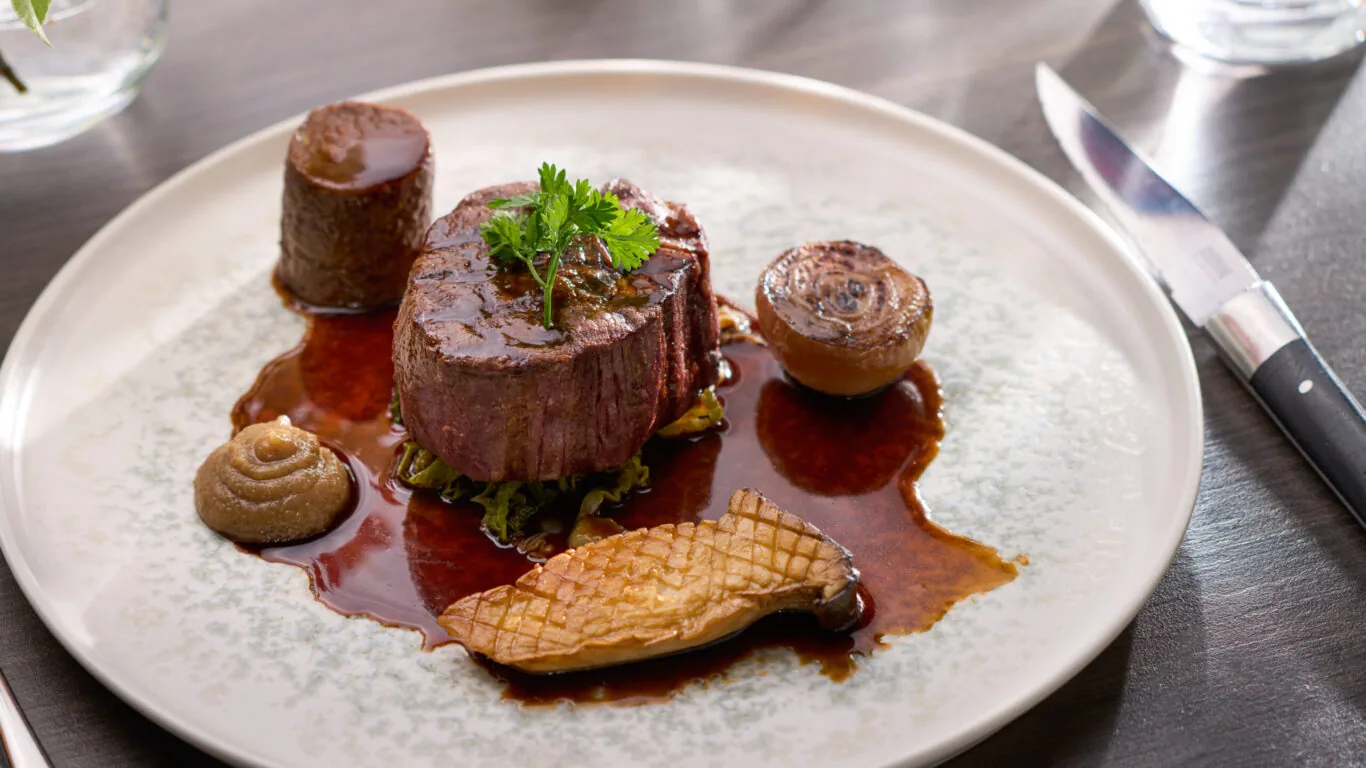 A plated gourmet dish featuring a filet of beef with sauce, a seared mushroom, an onion, a dollop of puree, and a crosshatched potato, garnished with fresh herbs.