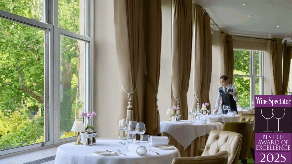 Elegant restaurant dining area with large windows, white tablecloths, and a waiter setting a table. 