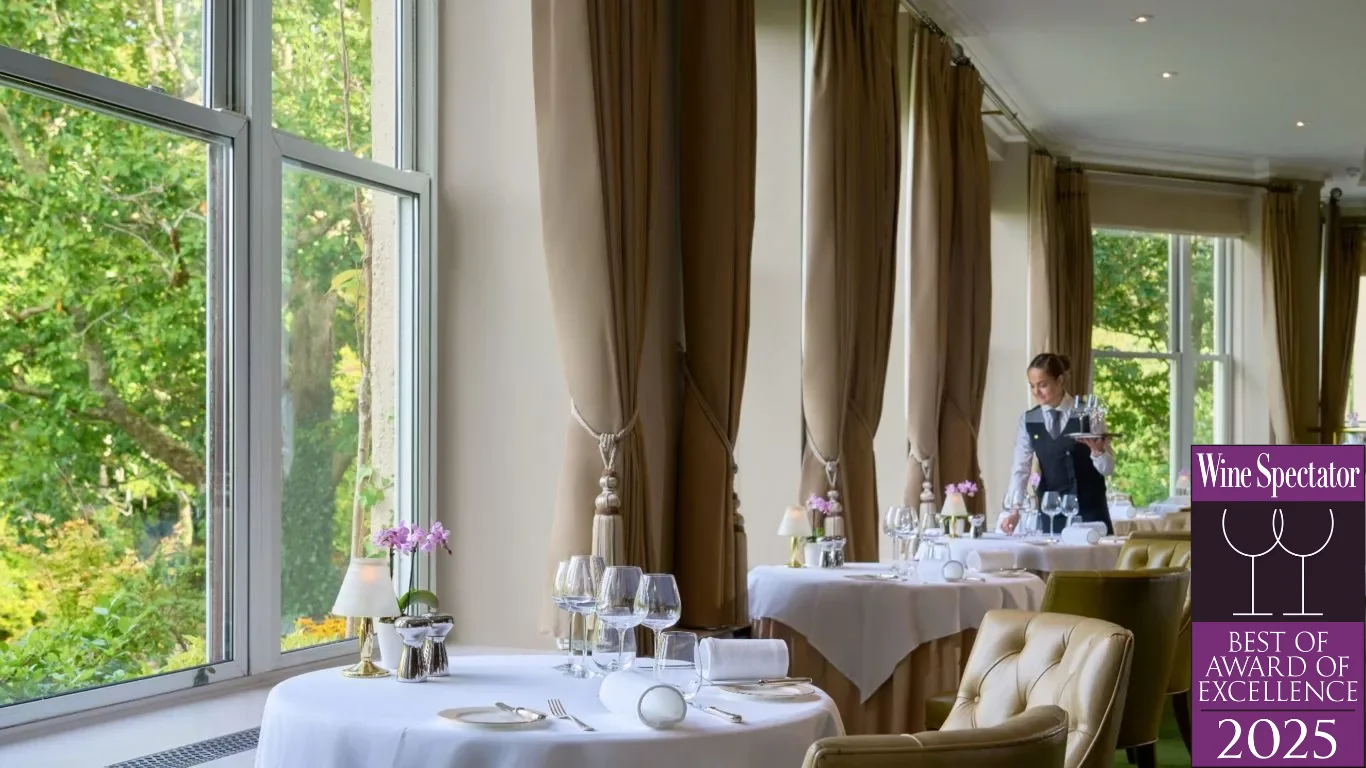Elegant restaurant dining area with large windows, white tablecloths, and a waiter setting a table. 
