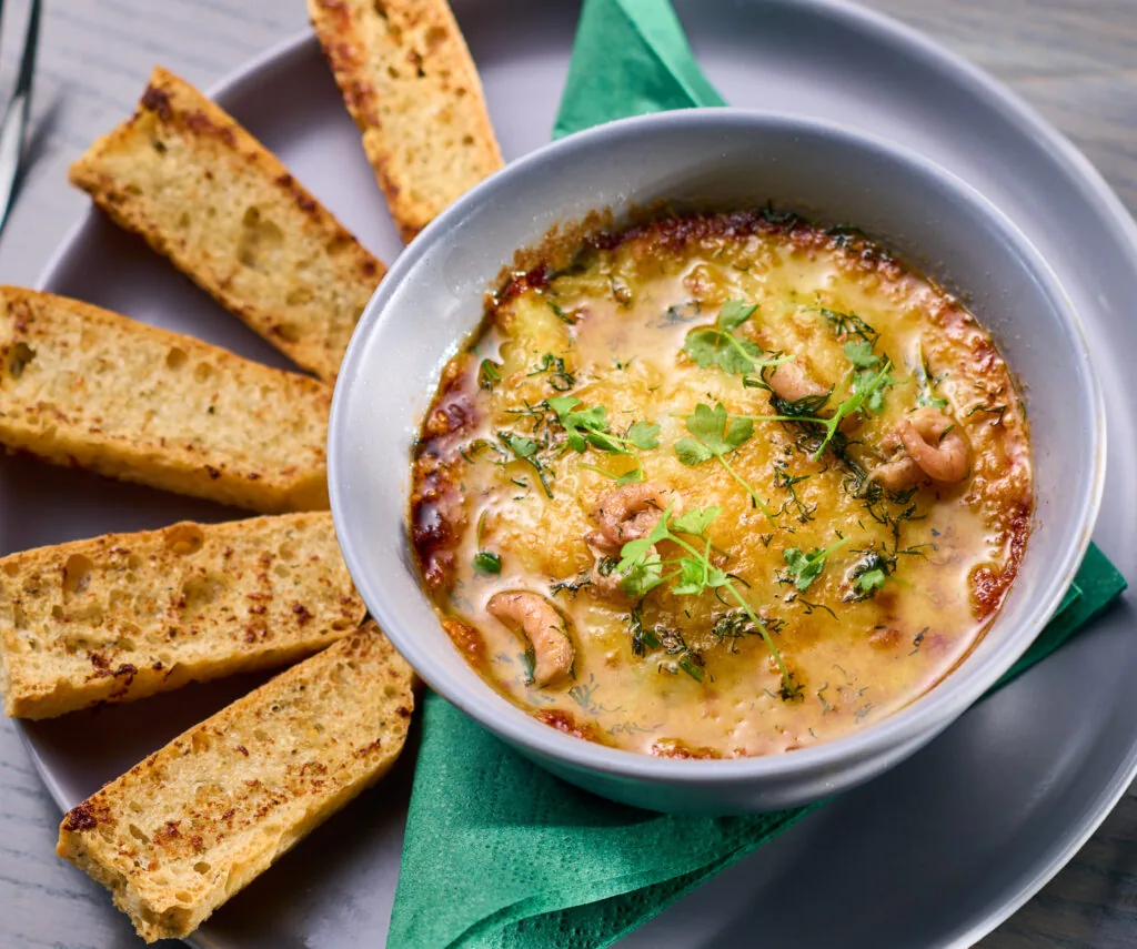 Bowl of baked cheese and nut dip garnished with herbs, served with several slices of toasted bread on a gray plate.