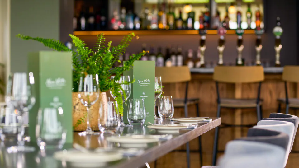 A modern restaurant table set with glassware, plates, and green menus, with a bar and barstools in the background.