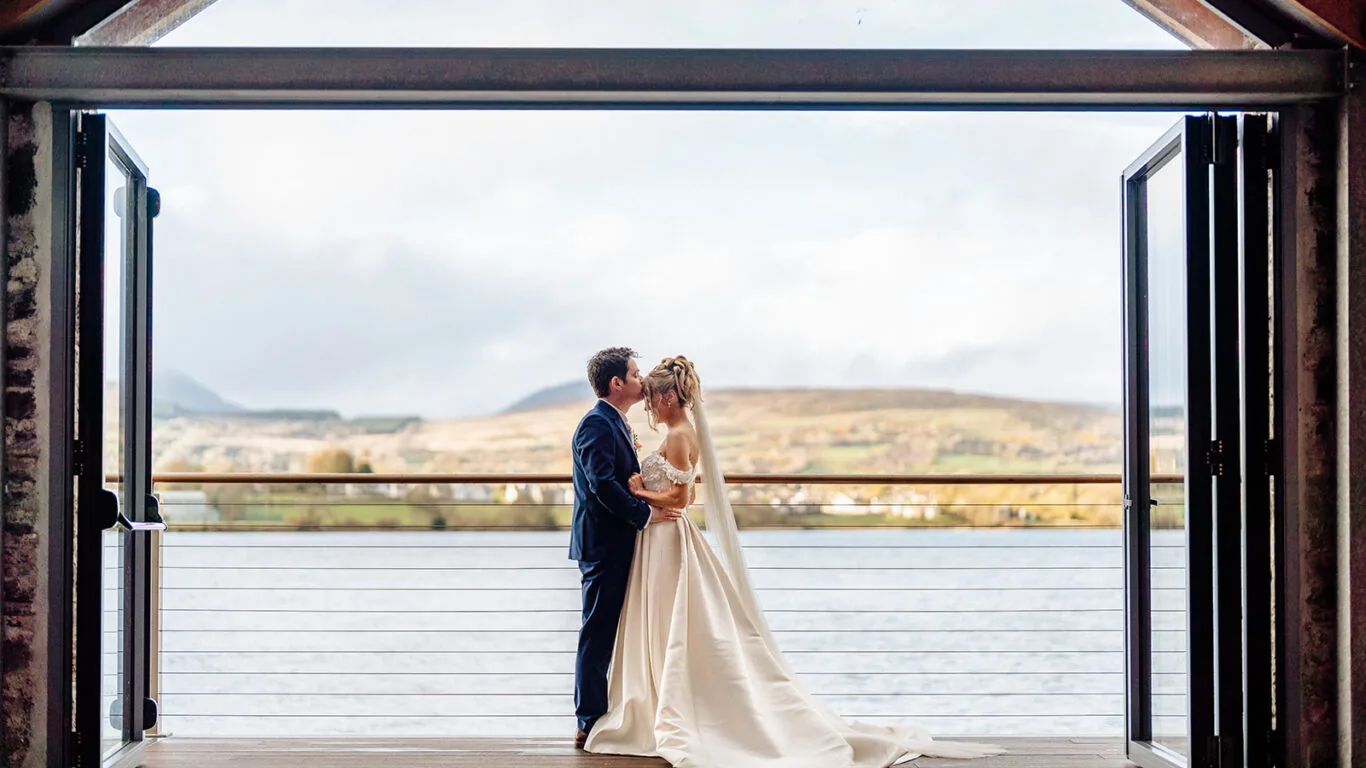 A bride and groom embrace and kiss on a wooden deck overlooking a lake and hills, framed by open doors.