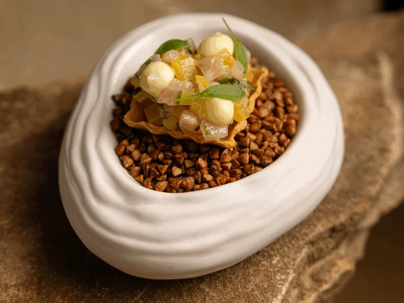 A small gourmet tart with diced toppings and cream dots is served in a white, oval dish filled with grains, set on a stone surface.