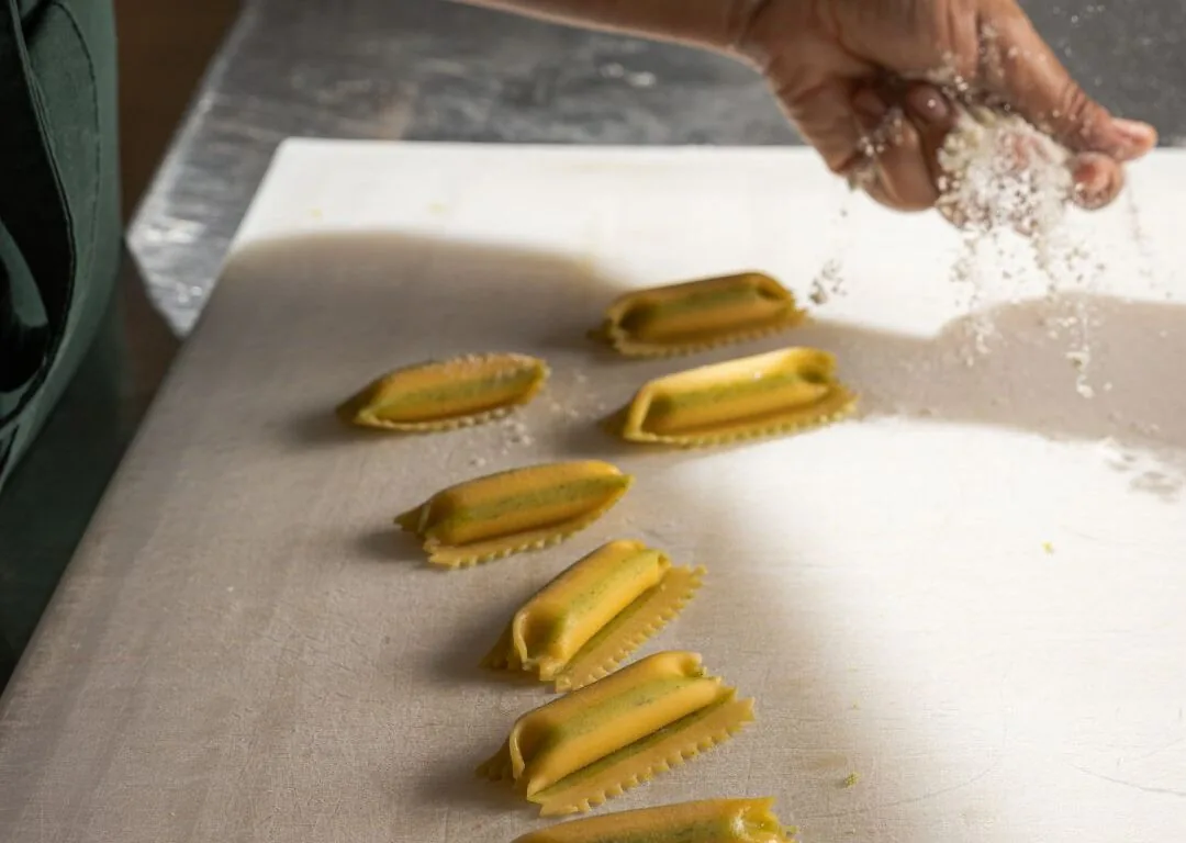 A person sprinkles flour over rows of uncooked stuffed pasta on a white cutting board.