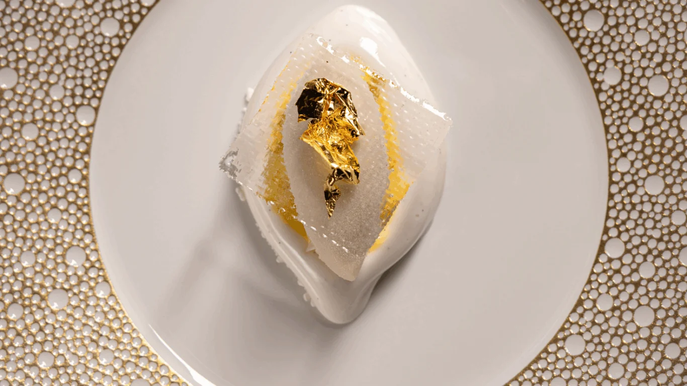 A plated dessert featuring a white glazed mousse topped with a thin gold leaf, served on a decorative white and gold-rimmed plate.