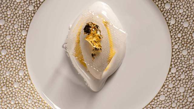 A plated dessert featuring a white glazed mousse topped with a thin gold leaf, served on a decorative white and gold-rimmed plate.