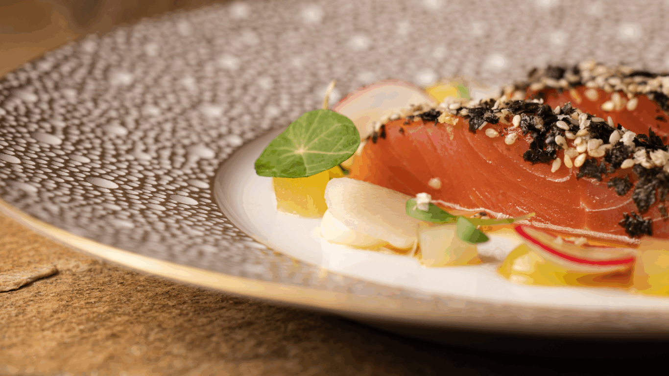 Slices of seared tuna coated with sesame seeds and herbs, garnished with radish, greens, and sauce on an ornate plate.