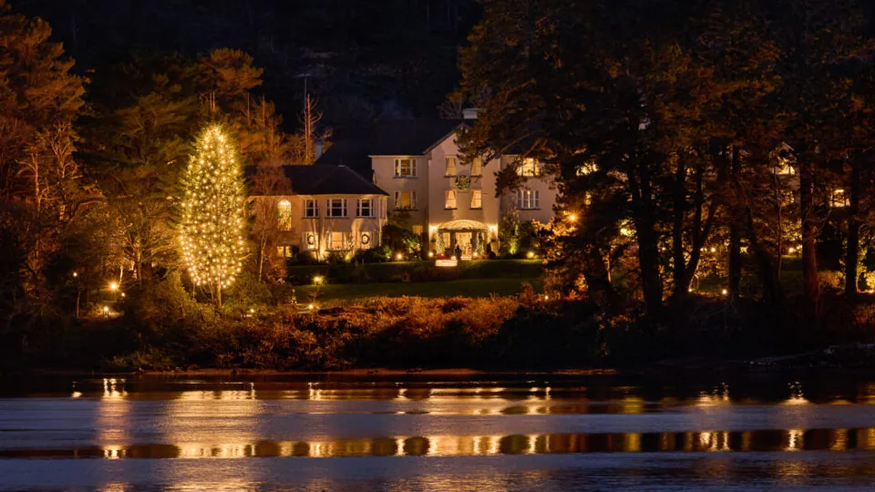 Sheen Falls Lodge surrounded by trees is illuminated with warm lights at night, reflected in the calm water of a lake in the foreground.