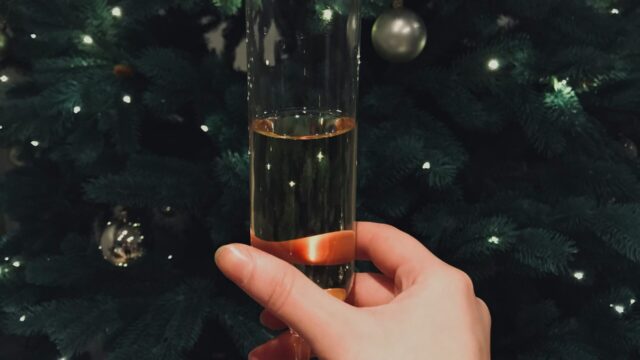 A hand holding a glass of champagne in front of a decorated Christmas tree with white lights and silver ornaments.