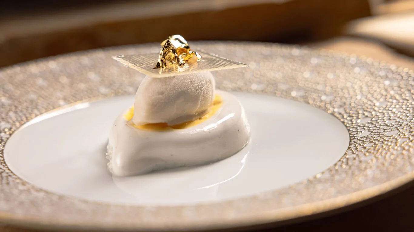A plated dessert features a scoop of white ice cream on a glossy white mousse, topped with a thin sugar sheet and edible gold leaf, served on an ornate plate.