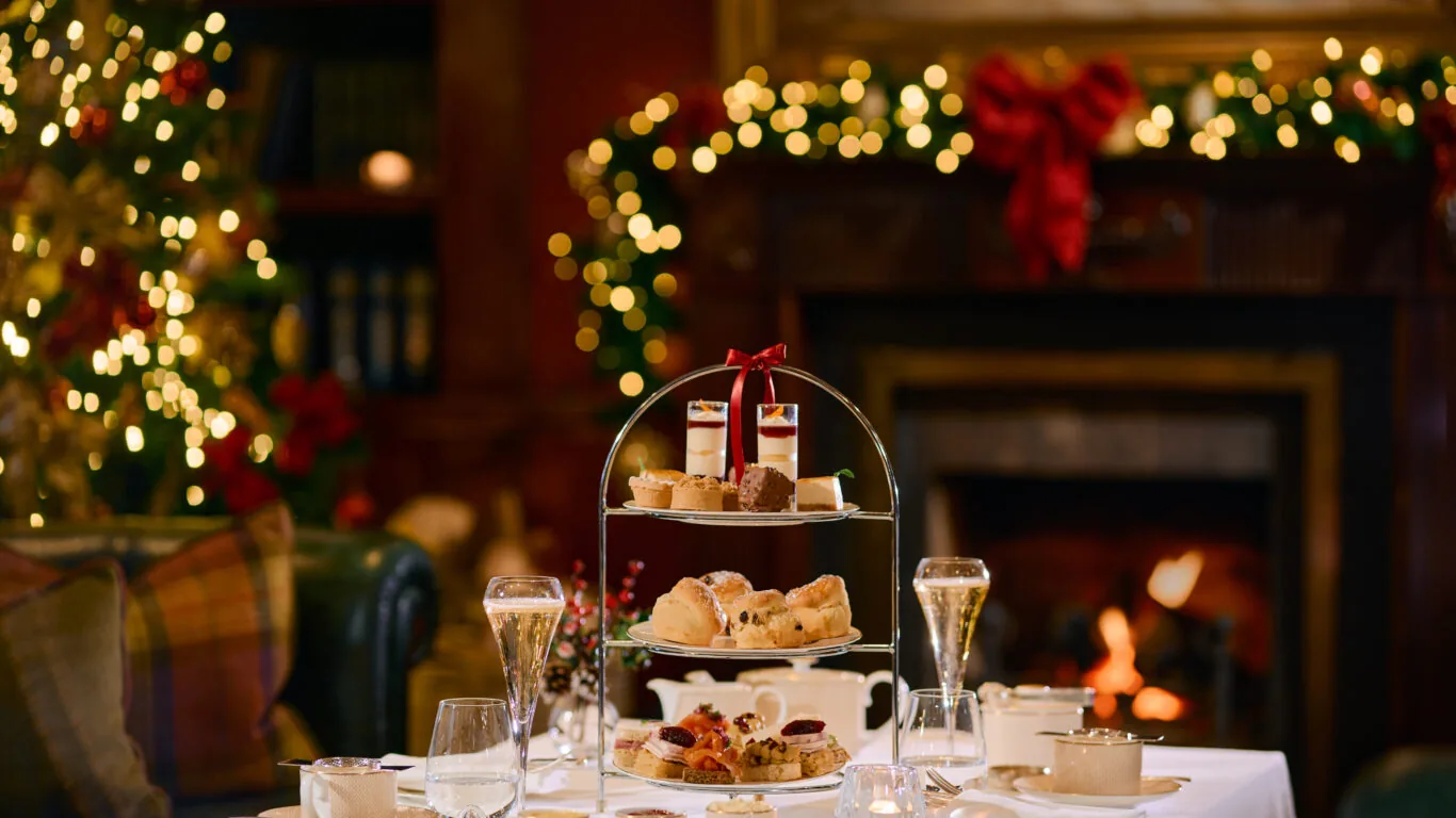 A festive afternoon tea setup with a tiered tray of pastries, scones, and sandwiches on a white tablecloth. A fireplace and decorated Christmas tree are in the background.