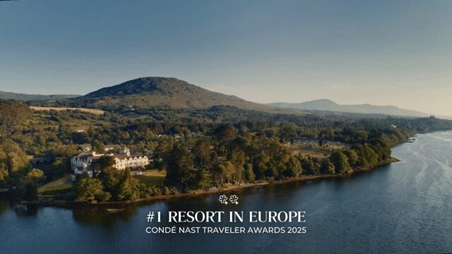 A large resort building surrounded by trees sits beside a lake with mountains in the background; text reads