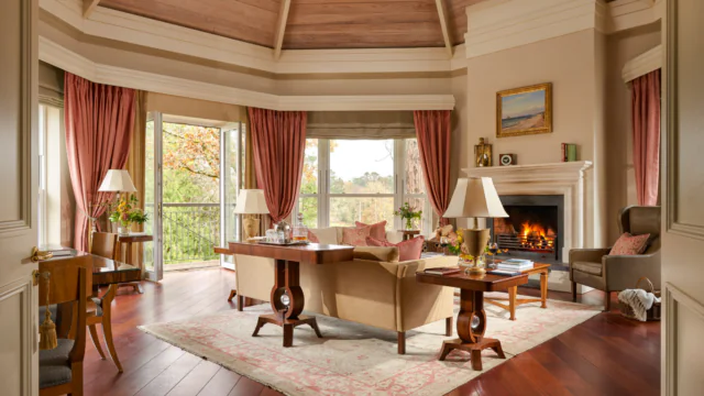A spacious, elegant living room features a fireplace, large windows with garden views, wooden floors, neutral furniture, and red curtains.