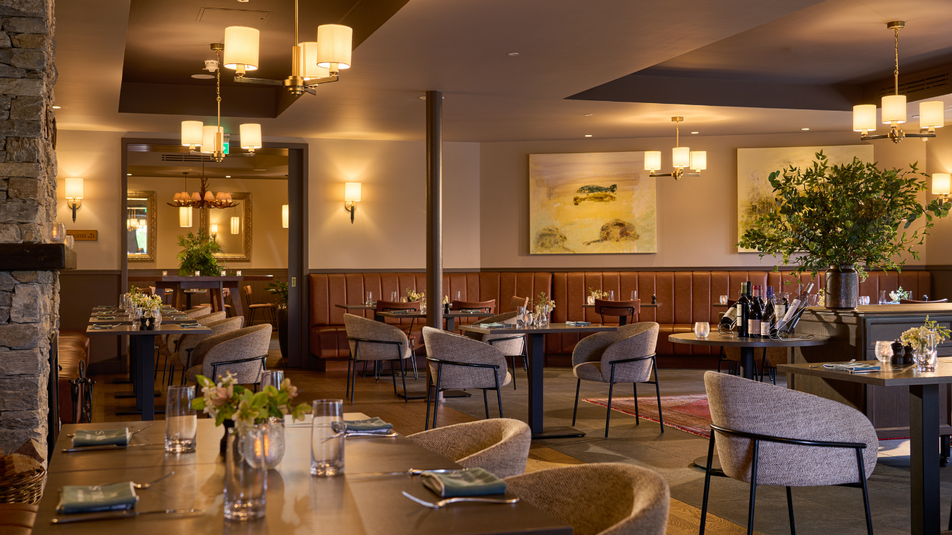 Modern restaurant interior with neatly arranged tables and chairs, warm lighting, wall art, and decorative plants, creating a welcoming dining atmosphere.