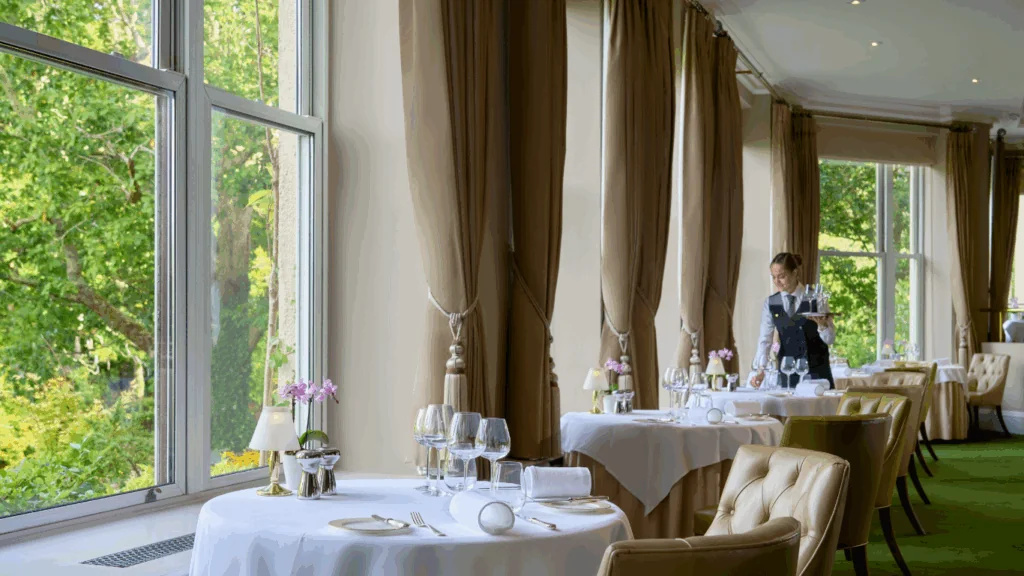 The Falls Restaurant A waiter sets a formal dining table in an elegant restaurant with large windows and views of green trees outside.