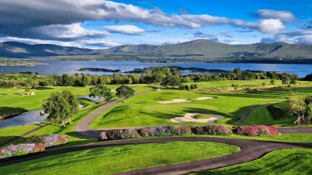 Golf A golf course with green fairways, sand bunkers, colorful flowerbeds, and a lake with mountains in the background under a partly cloudy sky.