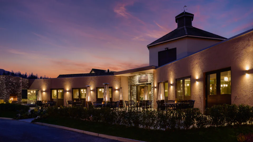 A modern building with outdoor seating and illuminated lights is shown at dusk under a colorful sky.