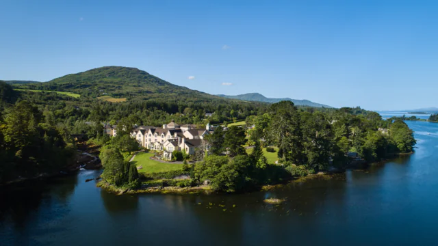 A large building surrounded by trees sits on a peninsula by a lake, with green hills and a blue sky in the background.