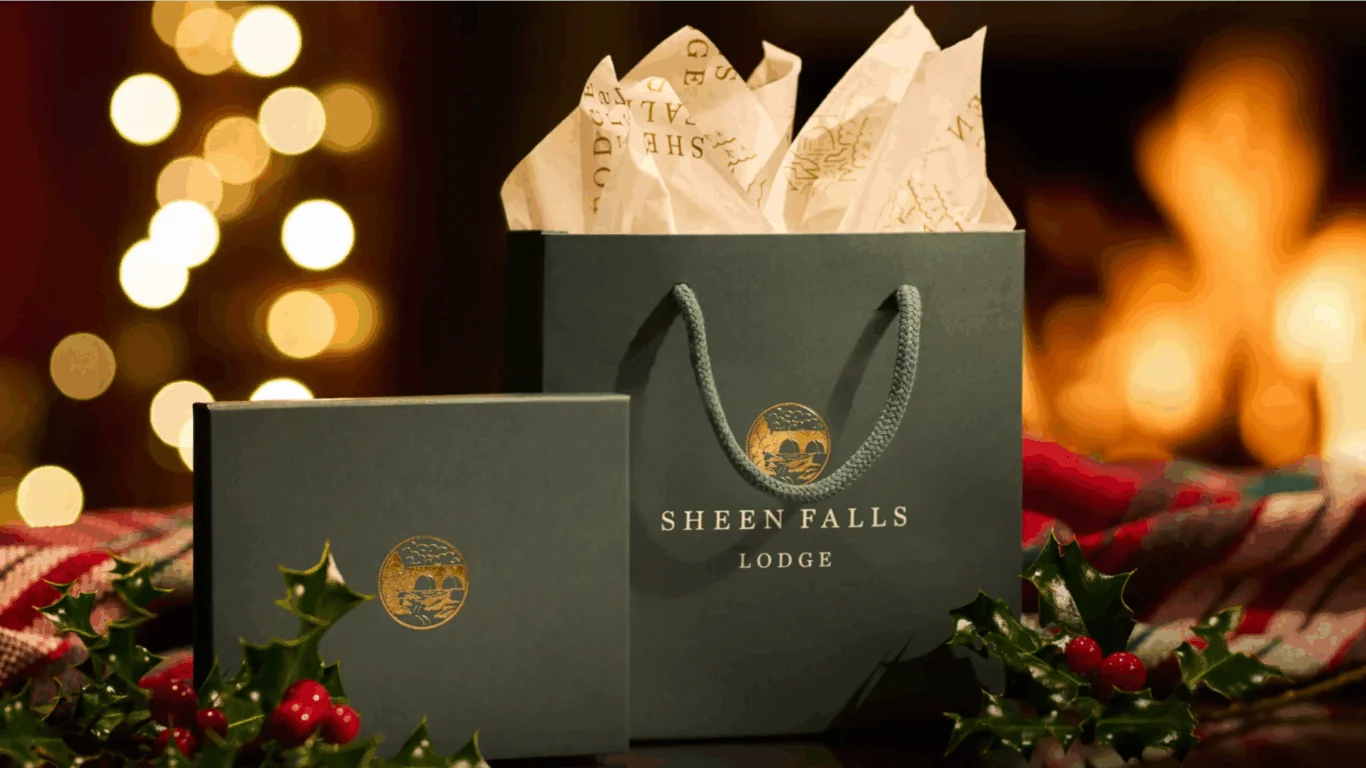 A Sheen Falls Lodge gift bag with tissue paper and a matching box, placed on a festive surface decorated with holly, with blurred lights in the background.