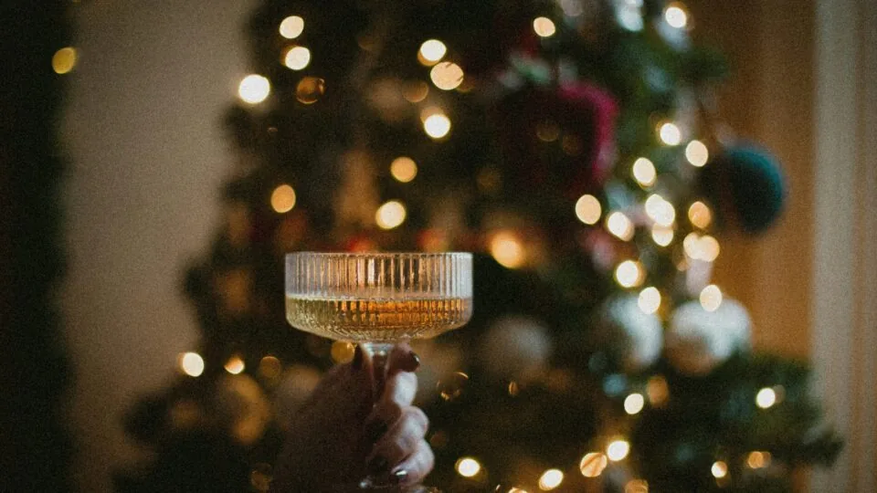 A hand holds a coupe glass of sparkling beverage in front of a decorated, lit Christmas tree.
