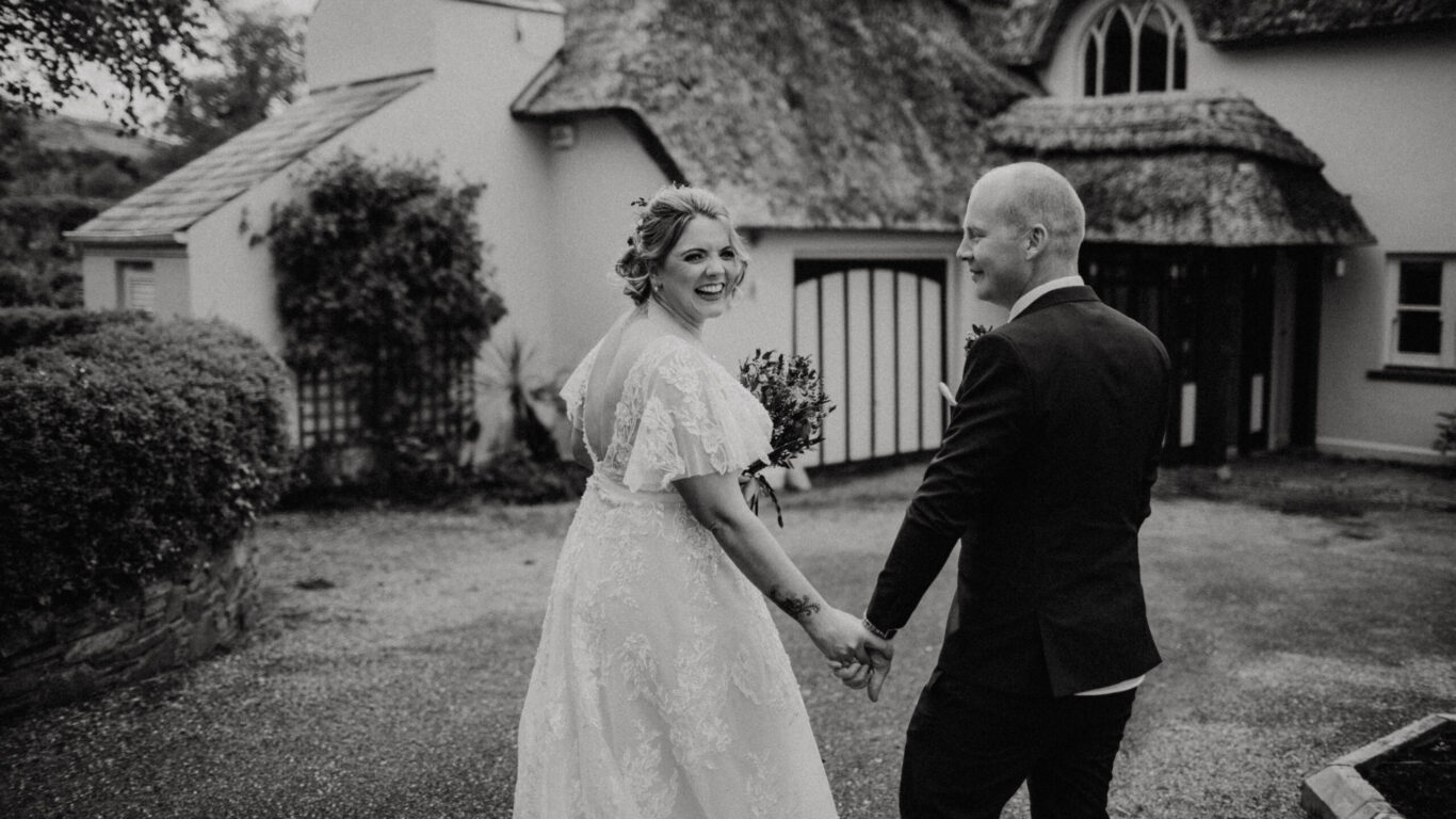 A bride and groom, holding hands and smiling, walk outside near a thatched-roof cottage. The bride holds a bouquet and wears a lace dress. The image is in black and white.